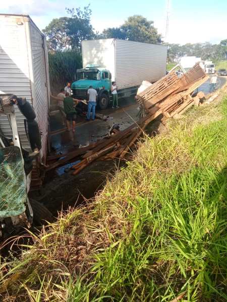 Acidente de caminhões na estrada, com carga de madeira caída e pessoas trabalhando para remover os destroços.