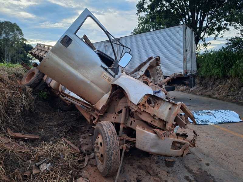 Caminhão acidentado na beira da estrada, parcialmente virado, com a cabine danificada e um caminhão ao fundo.