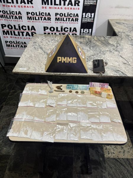 Mesa com itens apreendidos, incluindo pacotes transparentes, dinheiro em notas e moedas, e um objeto preto. Fundo com logo da Polícia Militar de Minas Gerais.