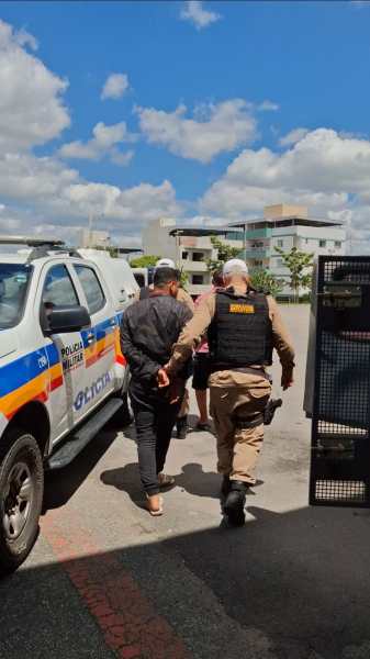 Policial acompanha suspeito algemado em área urbana, com viatura policial ao fundo.