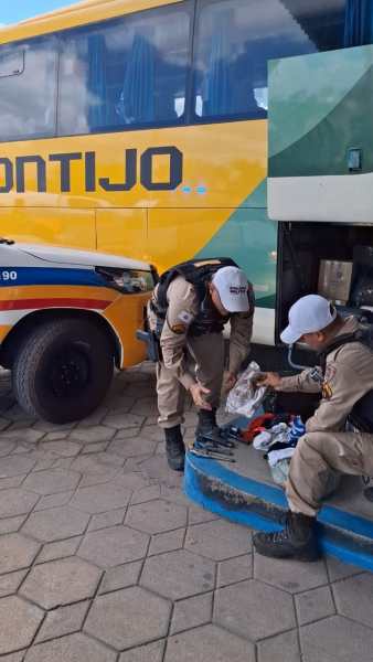 Dois agentes de segurança inspecionam roupas e outros itens em frente a um ônibus amarelo com a inscrição 'CONTIJO'.