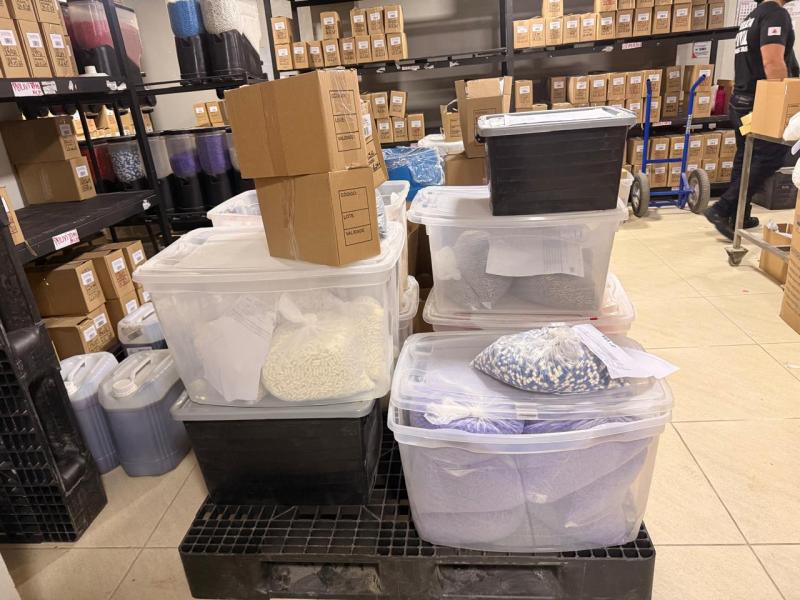 Imagem de um depósito com caixas empilhadas e recipientes plásticos transparentes cheios de materiais organizados. Prateleiras com diversos produtos etiquetados ao fundo.
