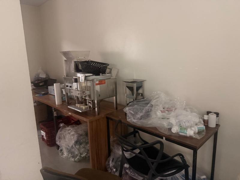 Ambiente com mesas e equipamentos de cozinha, incluindo um fogão e diversos utensílios plásticos e metálicos, cercados por materiais embalados.