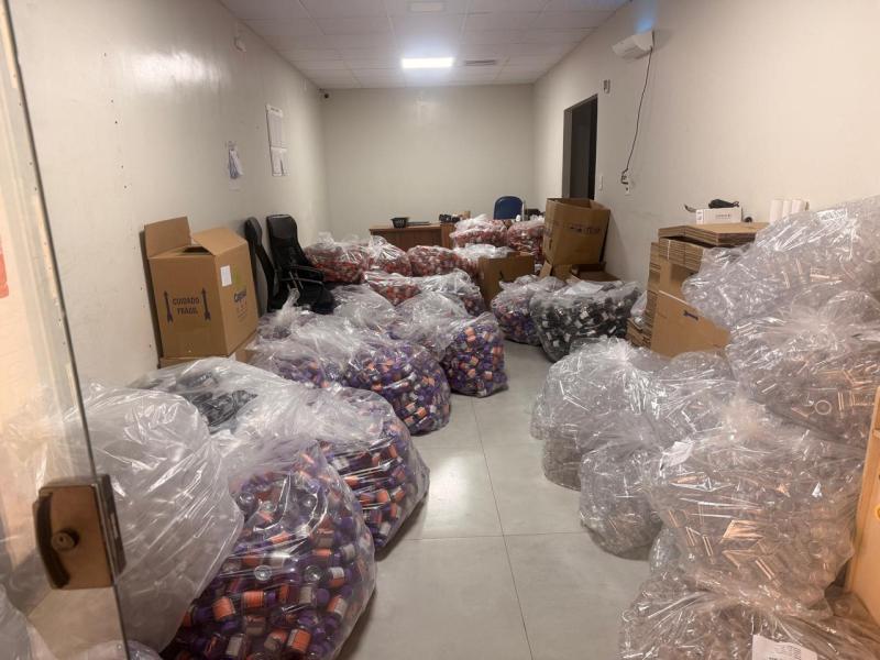 Sala com várias bolsas plásticas cheias de produtos, como frascos, dispostas no chão, ao lado de caixas de papelão e cadeiras.