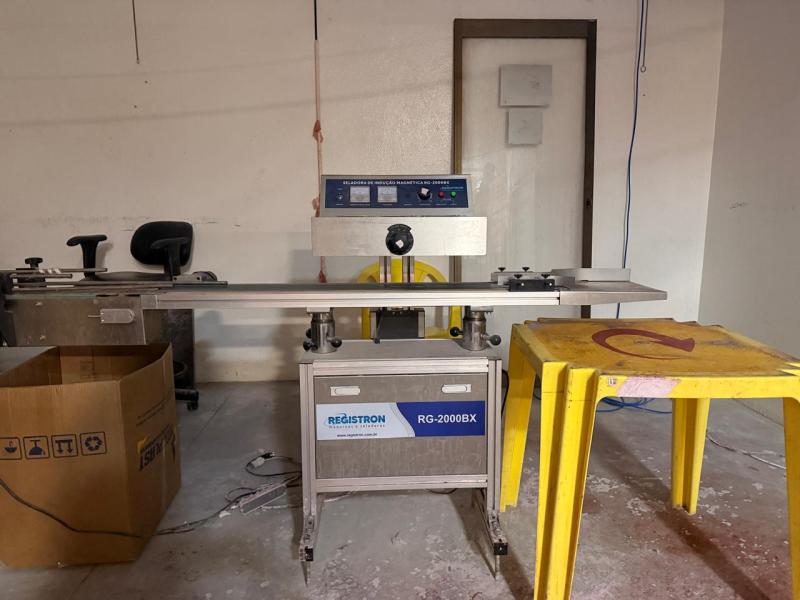 Máquina de pesagem e rotulagem modelo RG-2000BX em um ambiente de trabalho, com uma cadeira, uma caixa de papelão e uma mesa amarela no fundo.