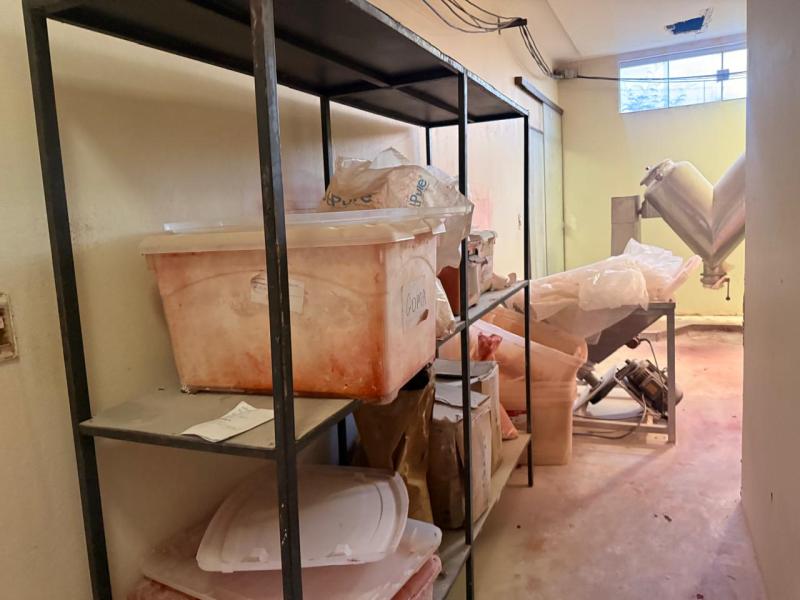 Interior de uma sala com prateleiras metálicas contendo caixas plásticas sujas e outros materiais embalados, com um equipamento de processamento visível ao fundo.