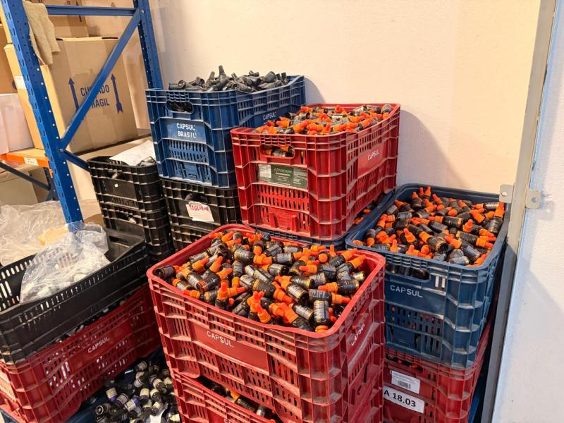 Caixas plásticas empilhadas contendo frascos com tampas laranjas em um ambiente de armazenamento.