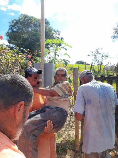 Um homem idoso sendo carregado por outra pessoa, enquanto duas outras pessoas observam. O cenário é ao ar livre, com árvores e flores ao fundo.