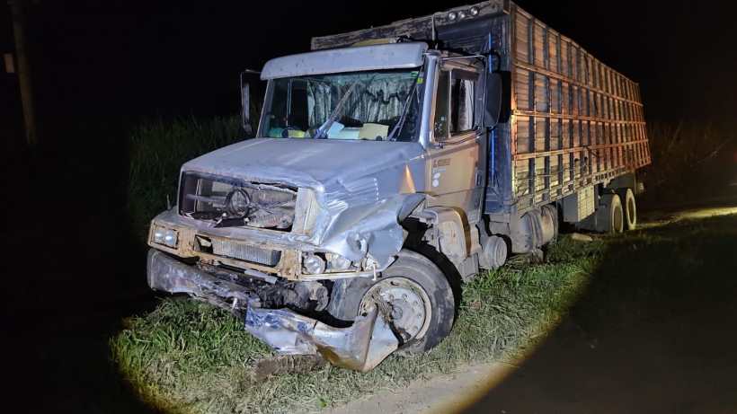 Caminhão danificado à noite, com parte frontal destruída, parado em estrada cercada por grama.