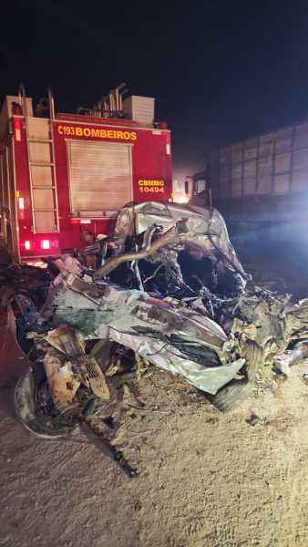Veículo destruído em um acidente, com partes retorcidas e queimadas, em frente a um caminhão dos bombeiros.