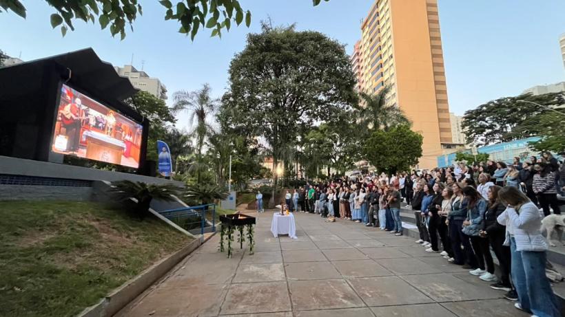 Praça com um grande telão exibindo uma apresentação, onde várias pessoas estão assistindo em frente a um altar com velas, cercadas por árvores e prédios ao fundo.