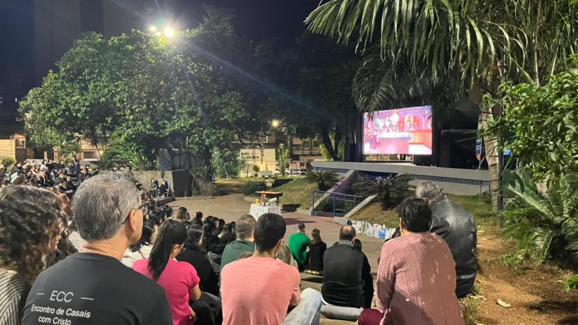 Grupo de pessoas assistindo a uma apresentação em um telão ao ar livre, com iluminação noturna e árvores ao redor.