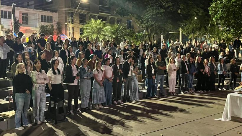 Grupo grande de pessoas assistindo a um evento ao ar livre à noite, com árvores e iluminação ao fundo.