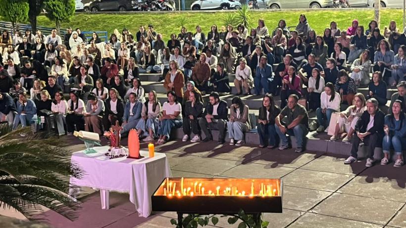 Uma multidão sentada em um espaço público, assistindo a um evento noturno, com uma mesa na frente decorada com velas e outros objetos.