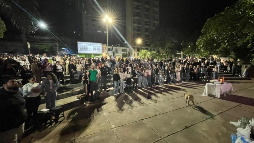 Pessoas reunidas em um evento noturno ao ar livre, com luzes e palmeiras ao fundo, algumas pessoas aplaudindo e um cachorro caminhando pelo local.