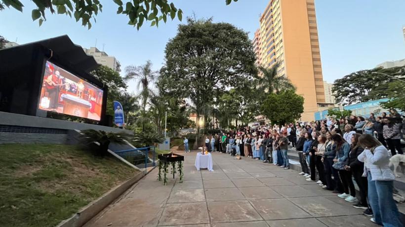 Evento ao ar livre com uma grande tela exibindo uma apresentação, enquanto um público de pé assiste em uma praça rodeada por árvores e prédios.