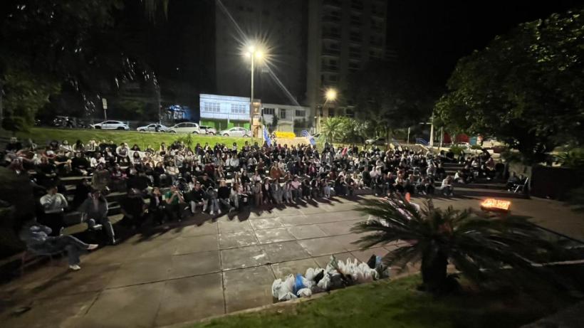 Grupo grande de pessoas sentado em um espaço ao ar livre à noite, com postes de luz iluminando a área e um edifício ao fundo.