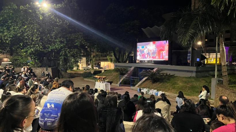 Pessoas assistindo a uma apresentação ao ar livre em um telão à noite, cercadas por árvores e iluminação.