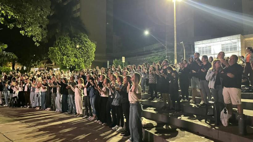 Pessoas aplaudindo em um evento noturno, em um espaço ao ar livre, rodeadas por árvores e edifícios.