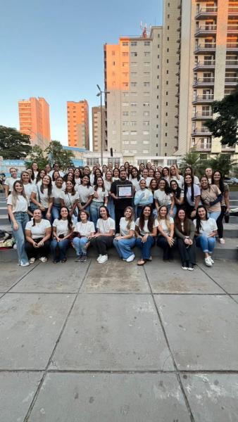 Grupo de mulheres sorridentes posando para foto em uma praça, com prédios ao fundo e segurando prêmios.