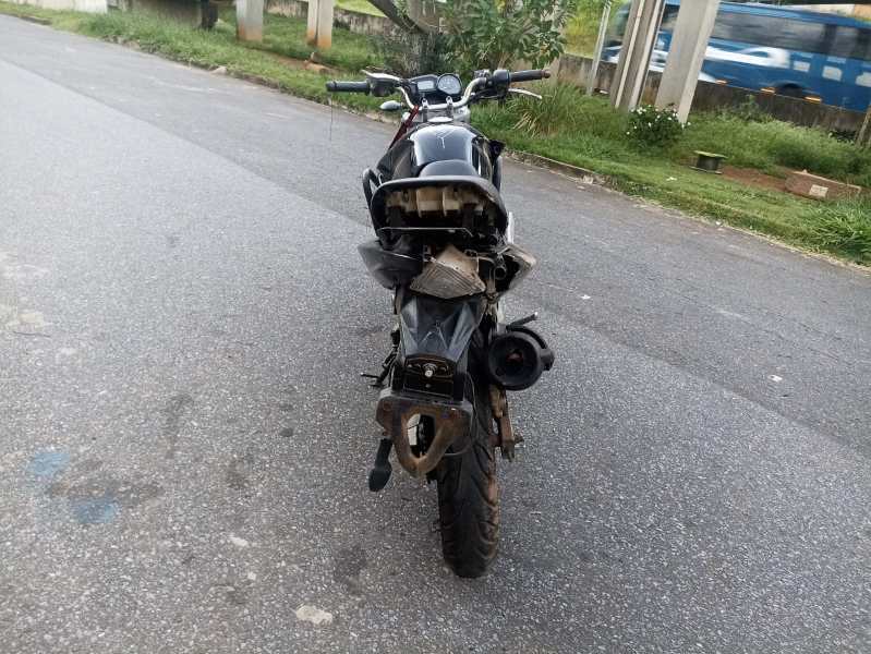 Motocicleta preta danificada vista de trás, estacionada em uma rua. O assento e a parte traseira estão visivelmente destruídos.