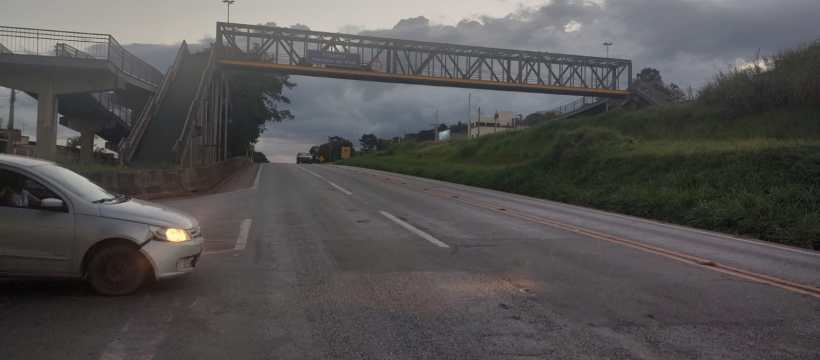 Um carro prateado está parado em uma estrada enquanto uma passarela metálica suspensa se encontra ao fundo, sob um céu nublado.