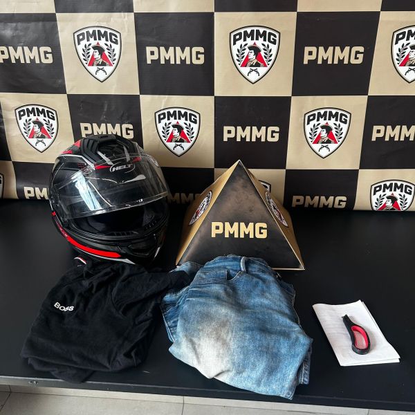 Um capacete, uma blusa preta, uma calça jeans e um pano branco sobre uma mesa, com um fundo de estande da PMMG.