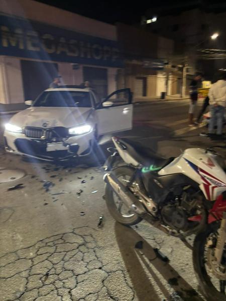 Cena noturna de um acidente envolvendo um carro e uma motocicleta na rua, com destroços visíveis.