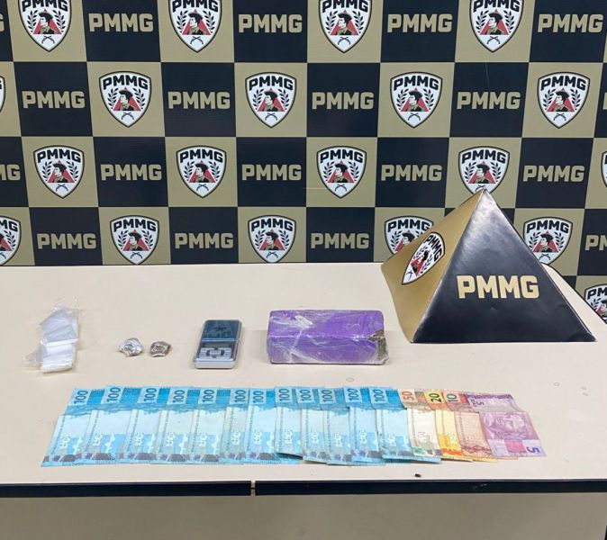 Mesa com notas de dinheiro, um bloco de substância, sacos plastificados e uma balança digital, com fundo de logotipo da PMMG.