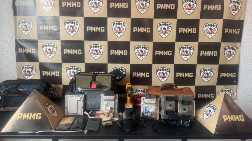 Mesa com ferramentas e equipamentos diversos, incluindo sacolas e dispositivos eletrônicos, em frente a um painel da PMMG.