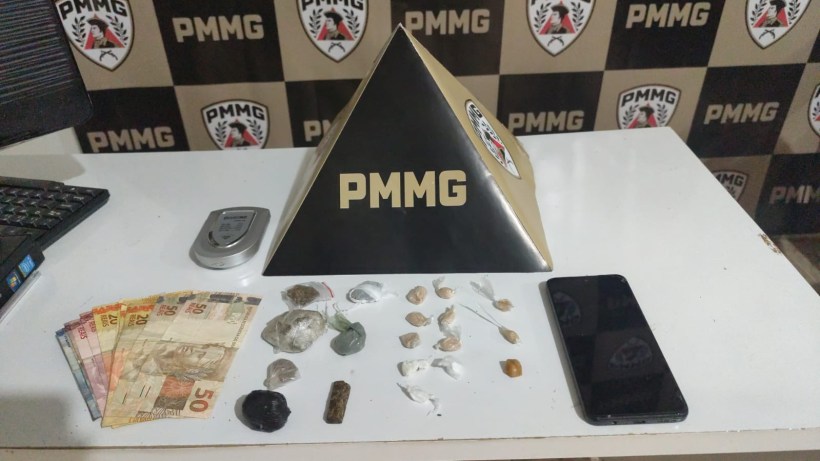 Uma mesa com uma pirâmide preta com a inscrição PMMG, um telefone celular, maços de dinheiro em reais e vários objetos, incluindo pedras e substâncias em pacotes.