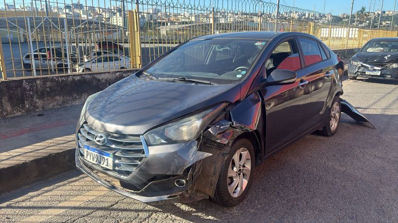 Carro preto batido com o lado esquerdo danificado, estacionado em uma área urbana.