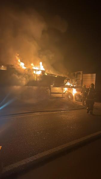 Caminhão em chamas à noite, rodeado de fumaça, com bombeiros tentando controlar o incêndio.
