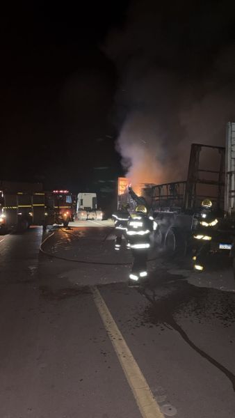 Bombeiros combatendo fogo em caminhão à noite, com fumaça visível e veículos de emergência ao fundo.