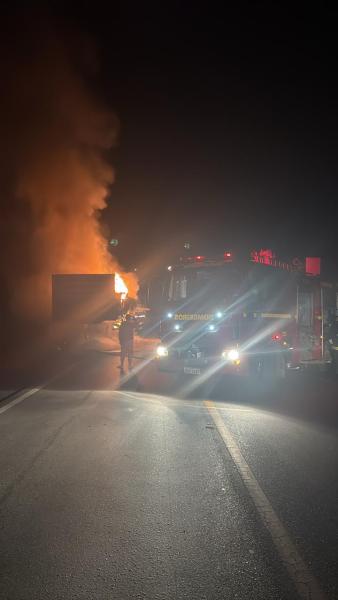 Caminhão de bombeiros atendendo a um incêndio noturno em um caminhão, com chamas visíveis e fumaça no ar.