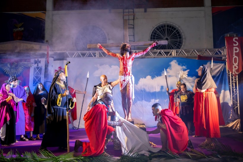 Recriação da crucificação de Jesus em uma encenação teatral, com personagens em trajes de época e um fundo dramático com fumaça e céu azul.