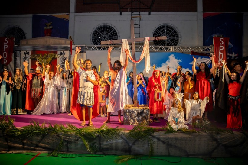 Cenas de uma apresentação teatral com diversos personagens vestidos em trajes históricos, incluindo uma cruz ao fundo e elementos de celebração.