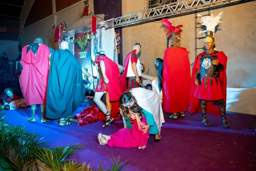 Grupo de pessoas participando de uma encenação teatral, vestindo roupas de época, com figuras históricas e um cenário de fundo, em um ambiente festivo.