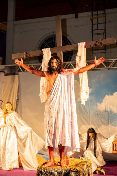 Cenário de encenação teatral com figura representando Jesus crucificado, braços abertos, vestido com roupas brancas e vermelhas, em meio a outros personagens com vestimentas brancas.