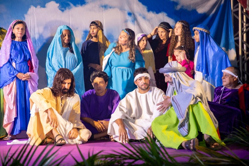Grupo de pessoas em trajes históricos em uma encenação teatral, com destaque para figuras centrais em poses contemplativas e outras ao fundo em trajes coloridos, representando um cenário bíblico.