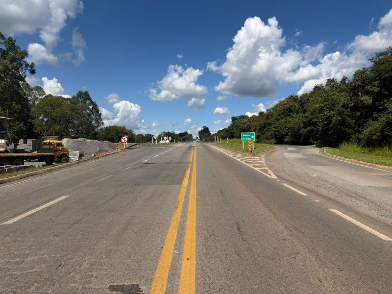 Estrada com pista dupla e faixas amarelas, cercada por vegetação e nuvens brancas no céu.
