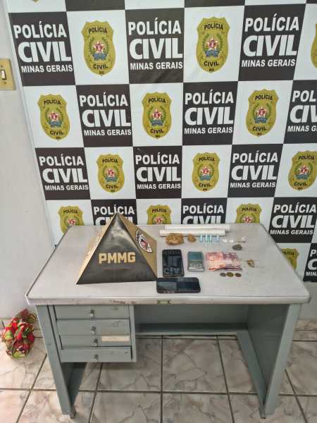 Mesa de trabalho com objetos apreendidos, incluindo celulares, moedas e uma pirâmide com a sigla PMMG, diante de um fundo com bandeiras da Polícia Civil de Minas Gerais.