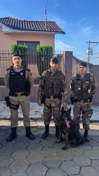 Três policiais em uniformes com um cachorro de polícia em uma rua residencial.