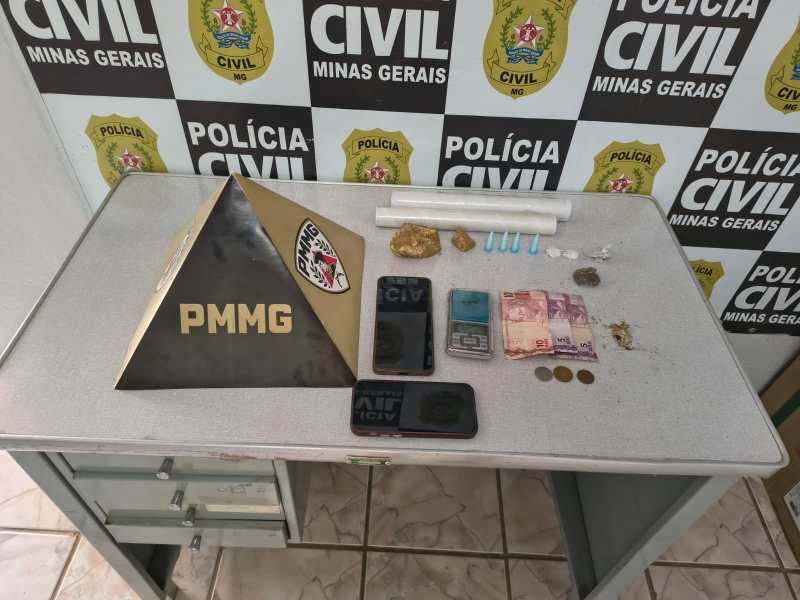 Imagem de uma mesa com uma placa da PMMG, dois celulares, notas de dinheiro, moedas, drogas e frascos, ao fundo um mural da Polícia Civil de Minas Gerais.