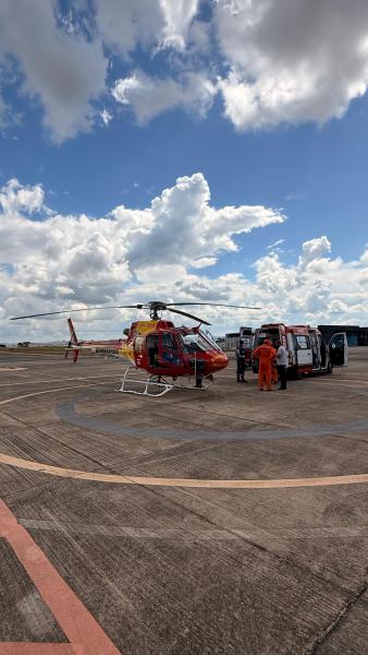 Helicóptero de resgate em um pátio, com equipe médica ao lado, em um dia ensolarado com nuvens.