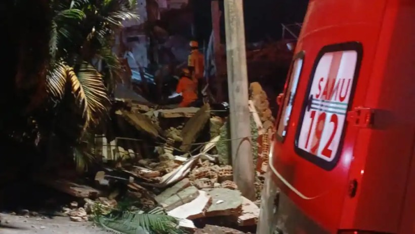 Cena de um lugar danificado, com escombros e uma ambulância do SAMU ao lado, enquanto equipes de resgate trabalham no local.