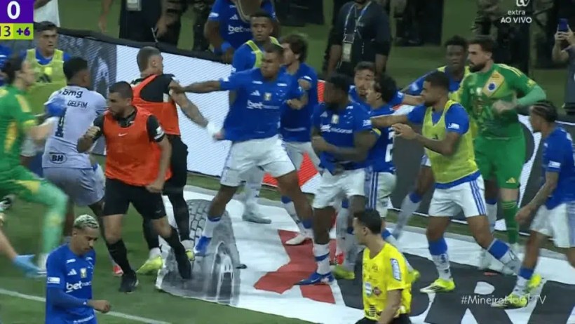 Jogadores de futebol de um time, uniformizados de azul, envolvidos em uma discussão acalorada durante uma partida, com um árbitro de camisa preta no centro da ação. O cenário é de tensão em campo, com a torcida ao fundo.