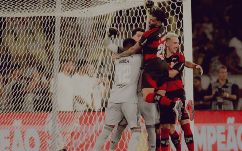 Jogadores de futebol comemorando um gol, se abraçando diante da rede do gol.