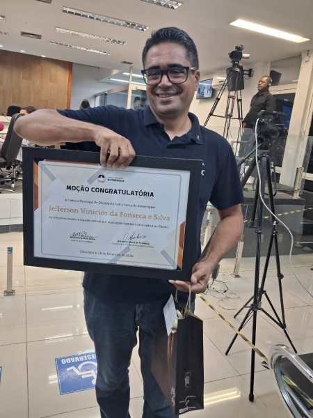 Homem sorrindo enquanto segura um certificado de moção congratulatória em um evento.