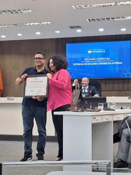 Cerimônia de entrega de moção de congratulações em reunião especial, com duas pessoas segurando um certificado em destaque e uma mesa com outros participantes ao fundo.